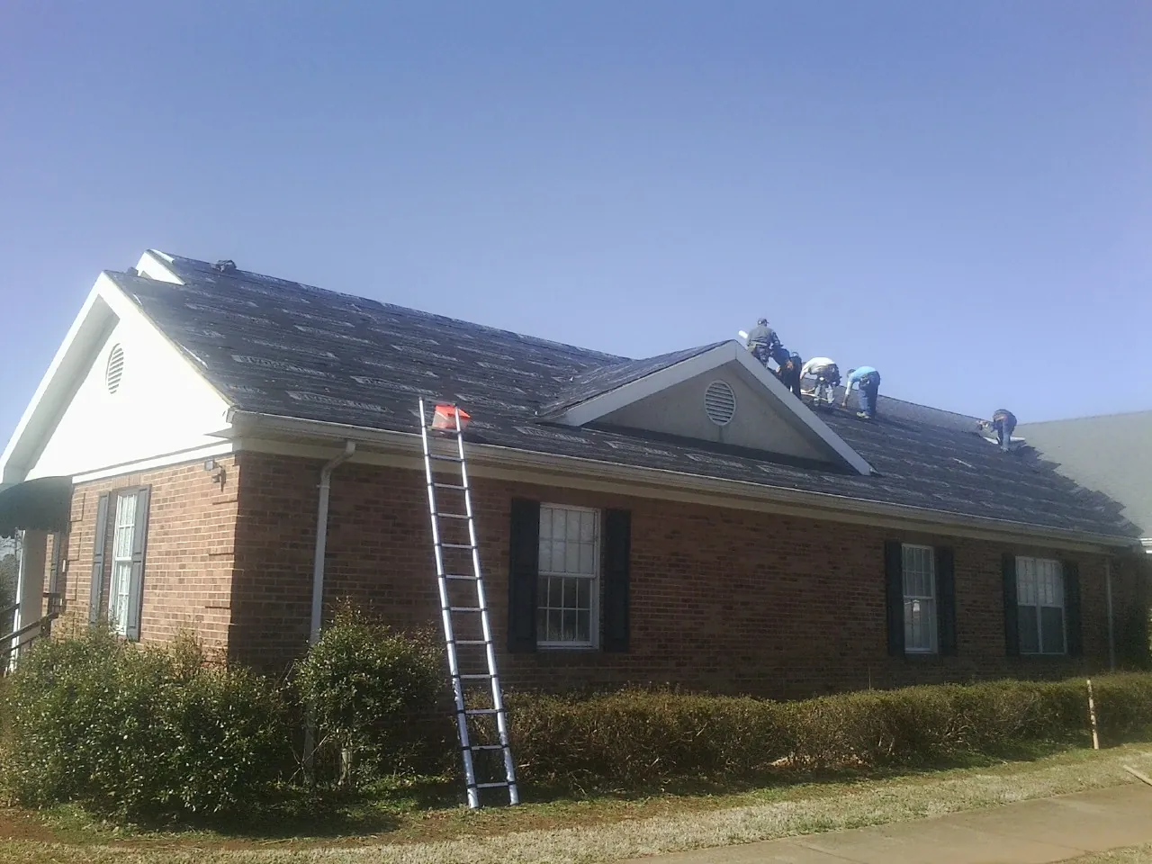 Crew tearing shingles off a brick ranch roof with a ladder set — Emergency Roof Repair work by our crew in Coleharbor