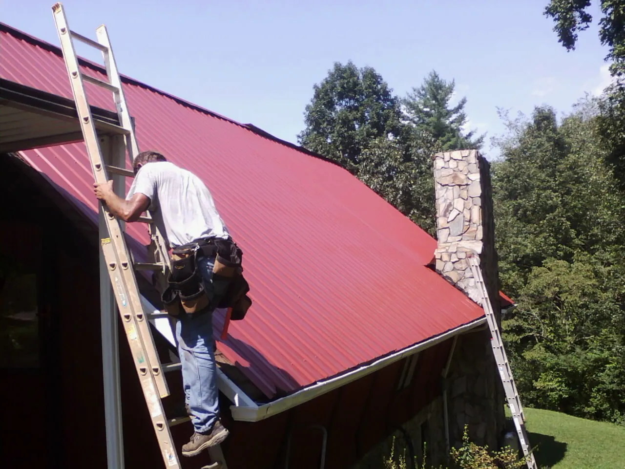 Roofer on a ladder beside a red metal roof and stone chimney — Soffit & Fascia Repair by Roofing Brothers in Coleharbor